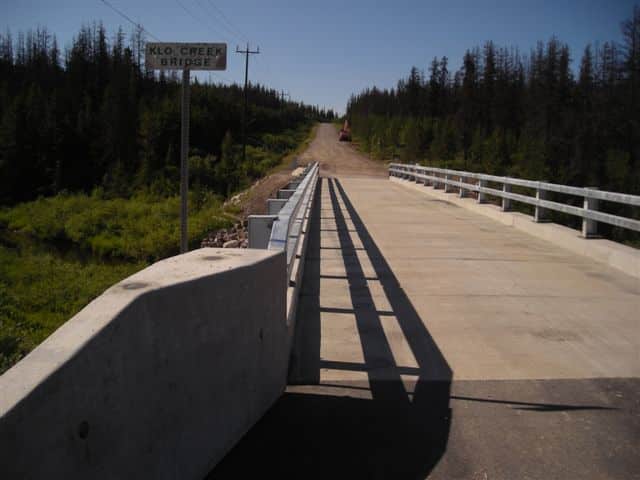 Concrete bridge labeled "Klo Creek Bridge" with metal railings, overlooking a dirt road and dense forest under a clear sky, exemplifies the commitment of leading highway bridge companies to infrastructure development.